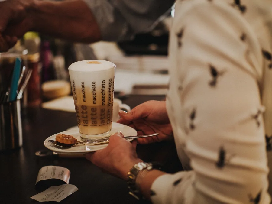 Ein Latte Macchiato im Glas wird mit einem Gebäckstück auf dem Unterteller über den Tresen gereicht