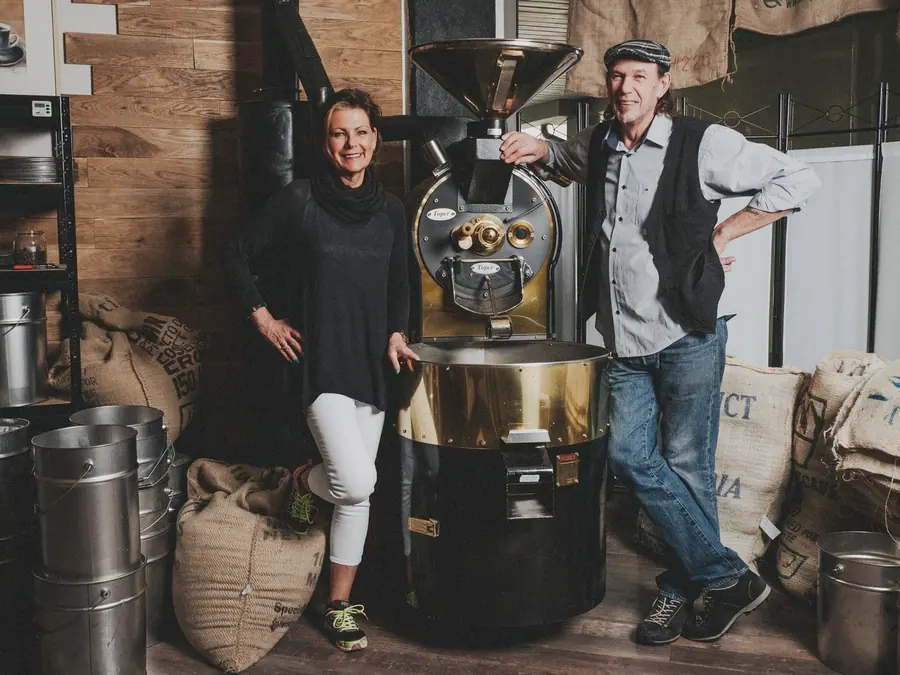 Sabine und Bernhard Schmitt stehen in ihrer Rösterei links und rechts neben dem Trommelröster, ringsum Jutesäcke mit Rohkaffee