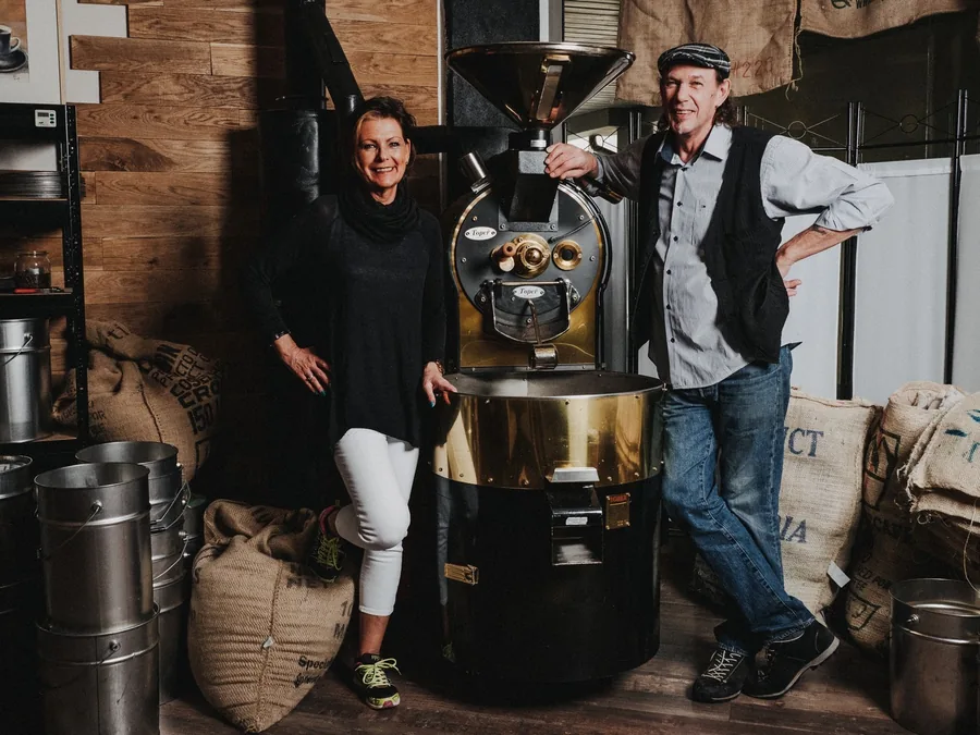 Sabine und Bernhard Schmitt stehen neben ihrem Trommelröster, ringsum Jutesäcke mit Rohkaffee